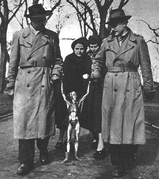 A black-and-white photo of small silver alien standing between two men dressed in trenchcoats and fedoras. The men are each holding onto one of the alien's hands, making it so the alien has its arms stretched far above its head.